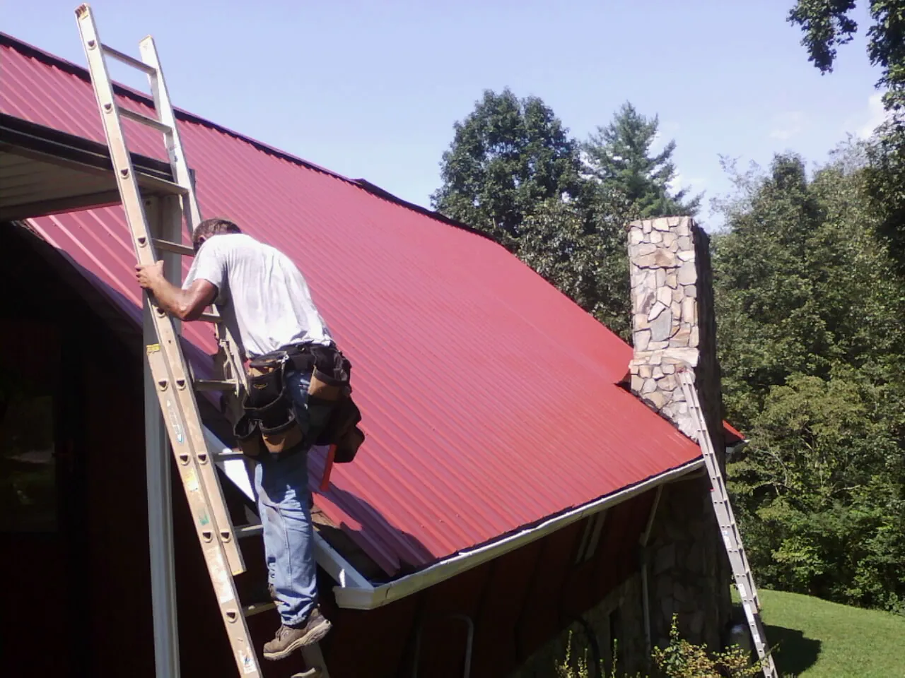 Roofer on a ladder beside a red metal roof and stone chimney — Soffit & Fascia Repair by Roofing Brothers in Gwynn Oak